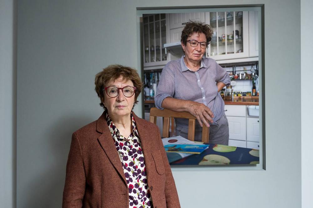 A woman in blouse and brown jacket stands in the exhibition in front of her portrait taken in a kitchen