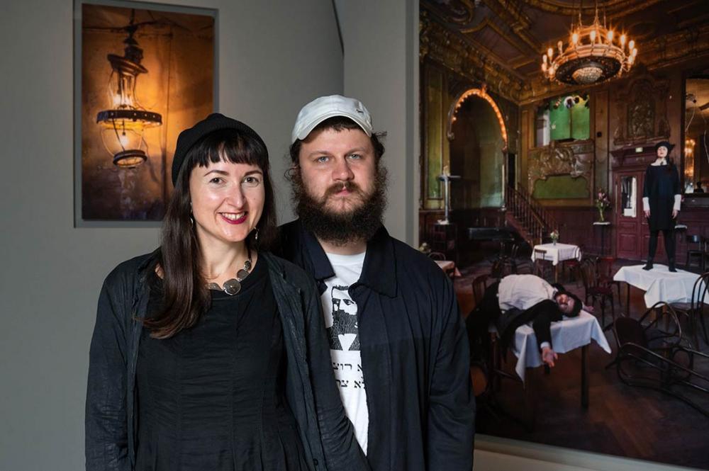 A laughing couple, she all in black, he in a black shirt and white baseball cap, stand in the exhibition in front of their portrait and the detail photo of a lamp from Clärchens Ballhaus 
