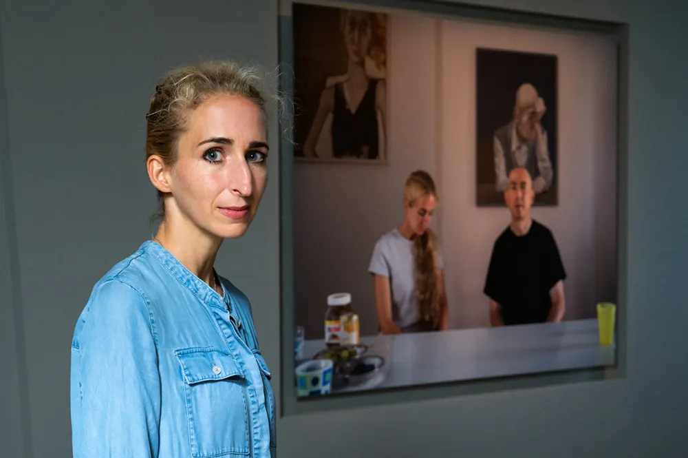 A blonde woman in a denim dress stands in the exhibition next to a photograph of herself and her partner sitting at the breakfast table, in the background two paintings on the wall