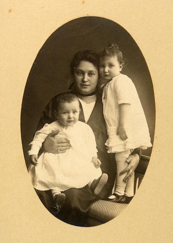 Ovale Studioaufnahme einer Frau, die ein Kleinkind auf dem Schoß hat, ein etwas größeres Kind steht neben ihr auf einem Stuhl