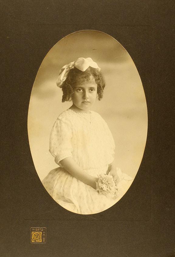Studioaufnahme eines Mädchens in weißem Kleid, mit Schleife im Haar und Stoffblume in der Hand