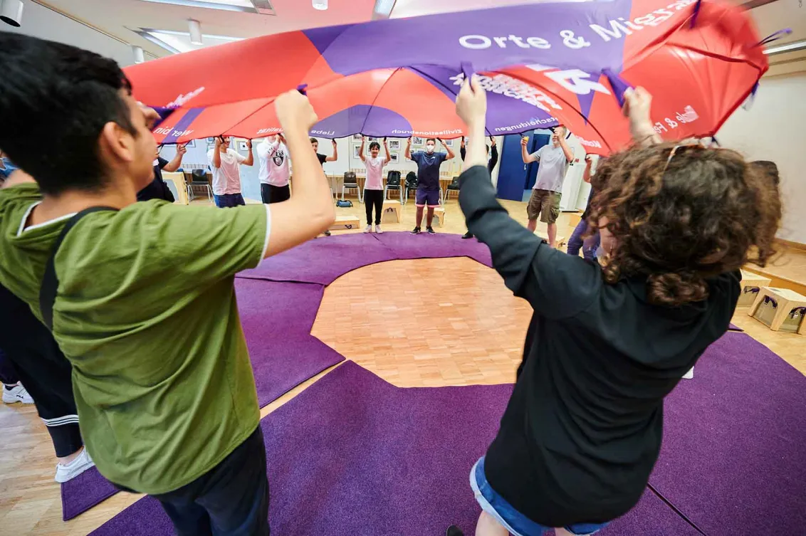A group of young people stand in a circle around a stretched-out parachute.