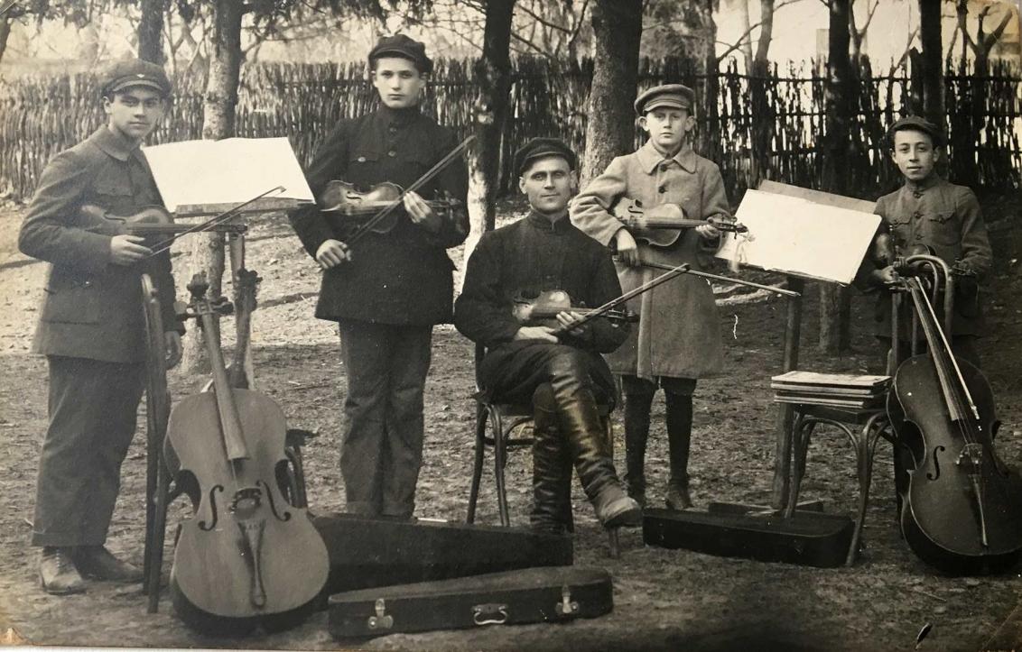 Black-and-white photograph of five young musicians with string instruments and music stands, outdoors