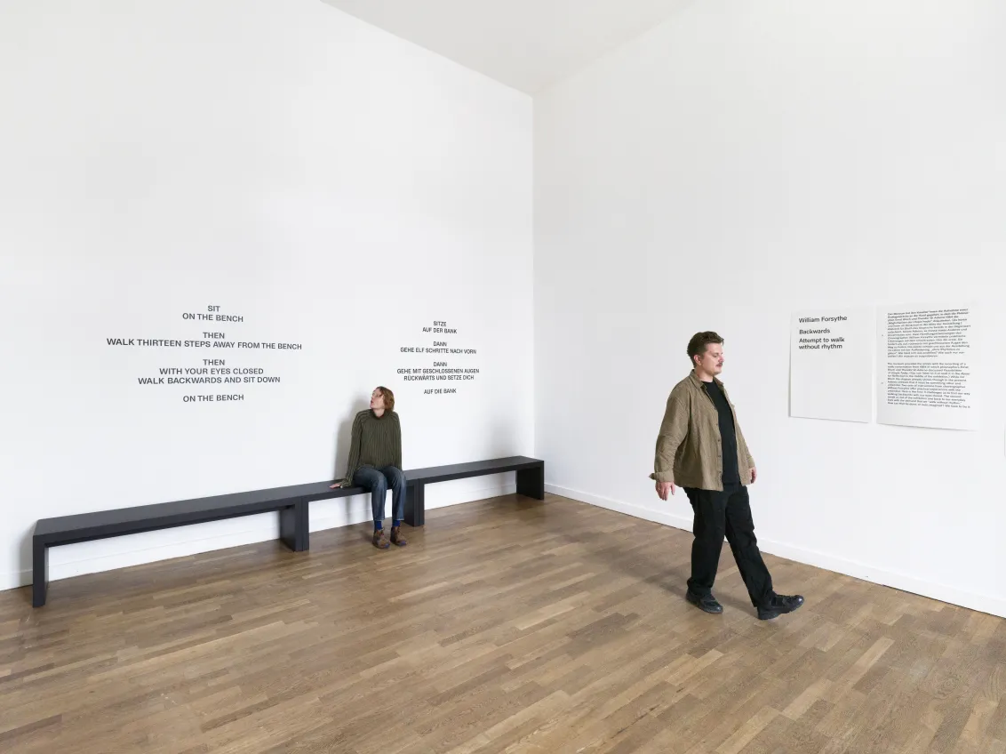 Eine Frau sitzt auf einer Bank. Ein Mann l&auml;uft r&uuml;ckwarts. Beide befinden sich in einem gro&szlig;en hellen Ausstellungsraum. 