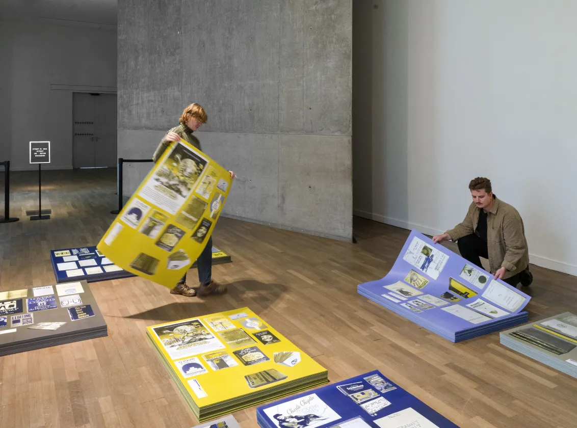Zwei Menschen nehmen gro&szlig;formatige beklebte Plakate von gro&szlig;en Stapeln, die auf dem Boden liegen in einem gro&szlig;en Ausstellungsraum.