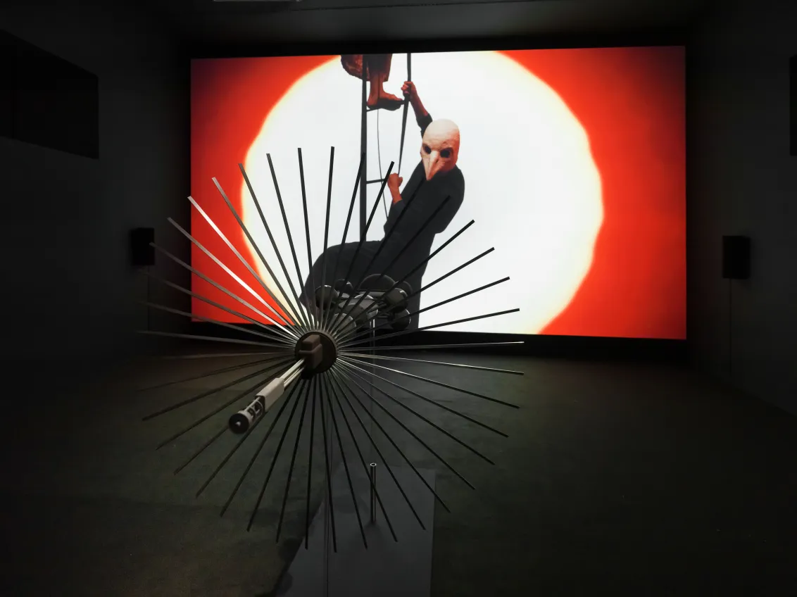 Futuristischer Metallgegenstand, der aussieht wie ein Ventilator, zeigt auf eine gro&szlig;e Filmleinwand. Auf der Leinwand steigen Personen eine Leiter empor. Eine Person hat eine Vogelkopfmaske auf. 