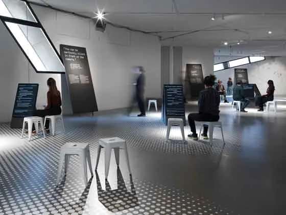 Das Bild zeigt einen Ausstellungsraum, in dem weiße Hocker verteilt sind, auf denen Menschen sitzen. Andere Menschen bewegen sich durch den Raum. Es gibt Aufsteller und Wände mit Text.