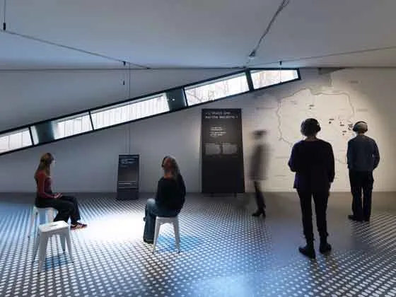 Das Bild zeigt einen Ausstellungsraum, in dem weiße Hocker verteilt sind, auf denen Menschen sitzen. Andere Menschen bewegen sich durch den Raum. Es gibt Aufsteller und Wände mit Text und einer Grafik..