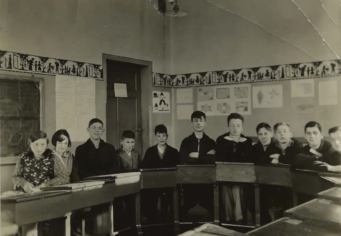 Das Bild zeigt einen Schulklassenraum mit elf Sch&uuml;lerinnen und Sch&uuml;lern, die in einem Halbkreis angeordnet an ihren Pulten sitzen.