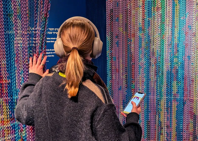 Portrait of a woman with long blonde hair and headphones in the Shabbat theme room. She is holding her mobile phone and following the JMB app Young Perspectives.