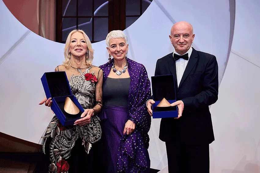 Drei Personen in festlicher Kleidung schauen freundlich in die Kamera. Zwei davon halten einen kupferfarbenen Preis in einer blauen Schatulle. 