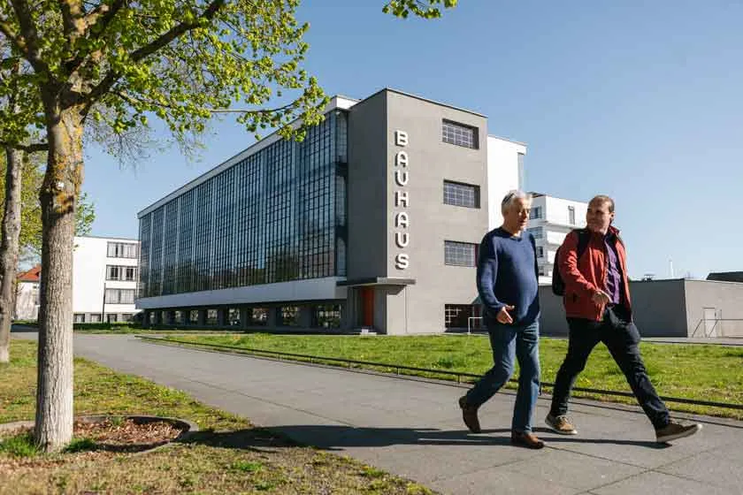 Zwei M&auml;nner laufen auf einem Weg nebeneinander. Hinter ihnen steht ein Haus mit der Aufschrift "Bauhaus".
