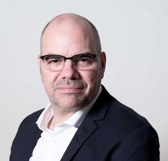 Ein Mann mit Halbglatze und Brille blickt direkt in die Kamera. er trägt ein weißes Hemd und einen schwarzen Sakko.