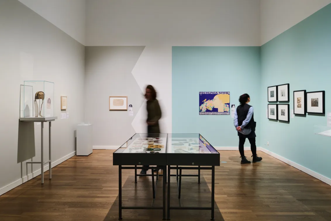 Ausstellungsansicht mit zwei Personen und Objekten, die in Vitrinen liegen oder an der Wand hängen.