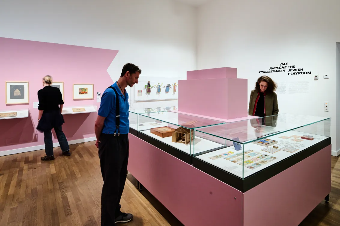 Ausstellungsansicht mit drei Personen und Objekten, die in Vitrinen liegen oder an der Wand hängen.