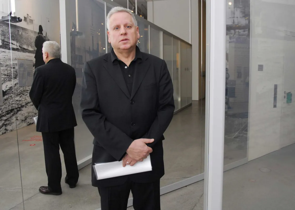 Stehender Mann in schwarzem Anzug vor einer verspiegelten Glasfläche, seine Körperrückseite ist als Spiegelbild zu sehen.