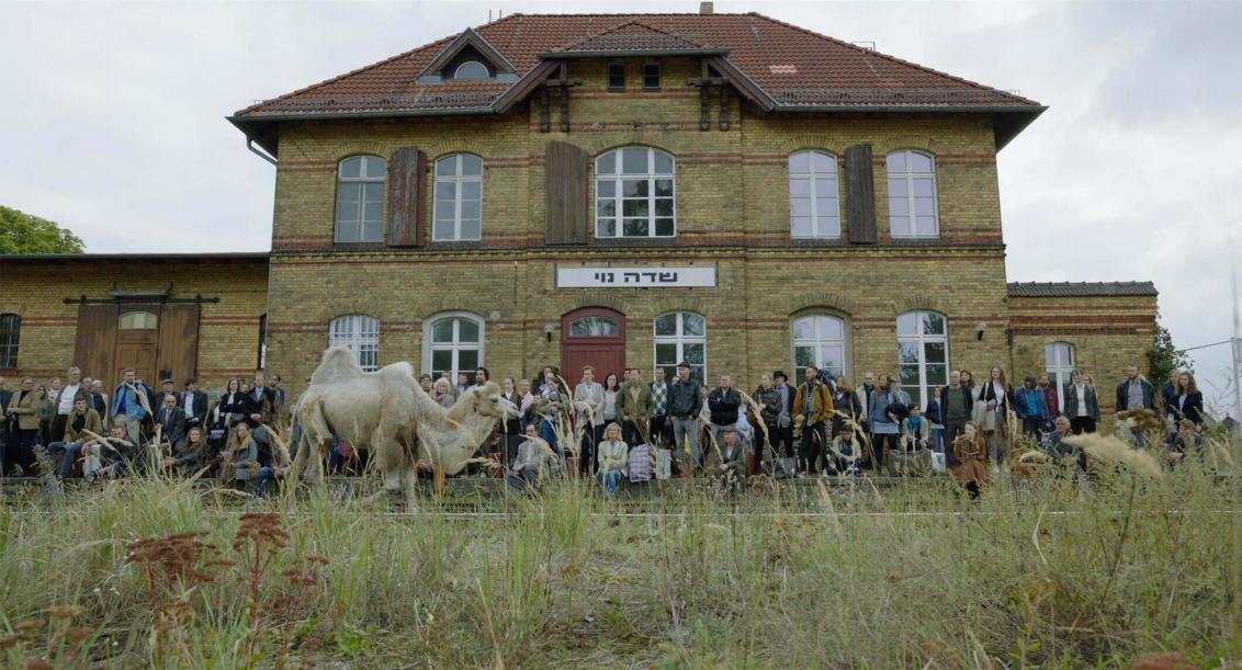 A larger group of people stands and sits in front of an old train station with a place name emblazoned on its facade in Hebrew script. In front of the waiting people, the railroad tracks and a camel following the tracks can be seen