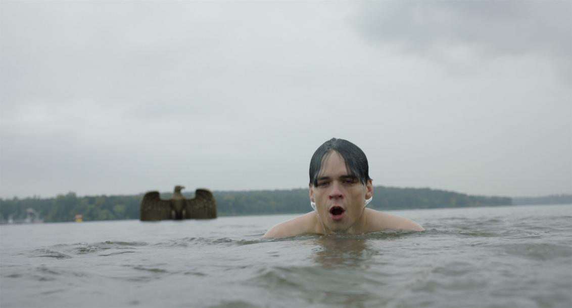 In the front of the picture you can see the wet head of a swimmer in a lake. Diagonally behind him, a large metal Imperial Eagle is just emerging from the water. The cloudy sky and the water surface are gray in gray and look threatening