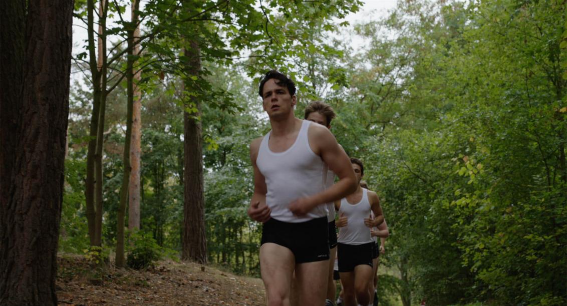 A group of young men in white jerseys and black, very short pants jog in single file through a summer-green deciduous forest