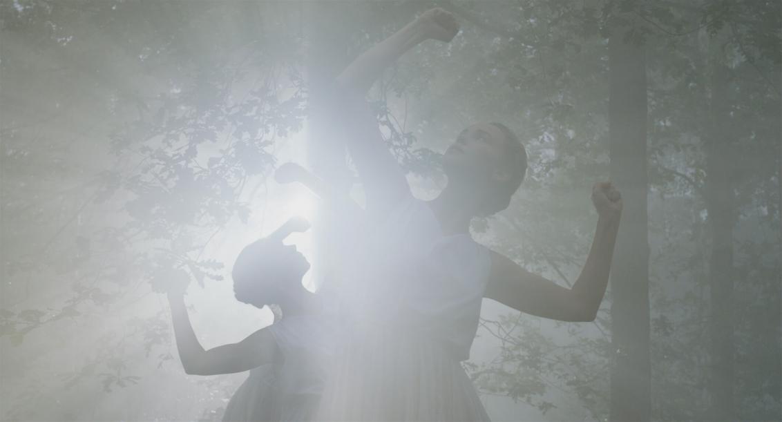 Junge Frauen in weißem ärmellosen Sommerkleidern tanzen in einem Wald im Gegenlicht. Ihre Oberarme bilden eine Linie mit ihren Schultern, die Unterarme zeigen in rechtem Winkel mit geballten Fäusten nach oben