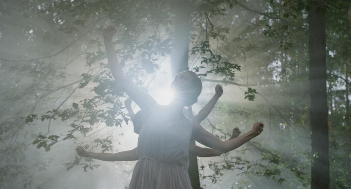 Junge Frauen in weißem ärmellosen Sommerkleidern tanzen in einem Wald im Gegenlicht. Sie recken ihre Arme zeigen mit geballten Fäusten