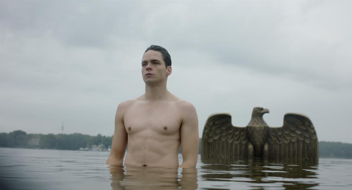 A man stands in a lake, his naked upper body and his head are visible. Behind him, a metal Imperial Eagle rises out of the water
