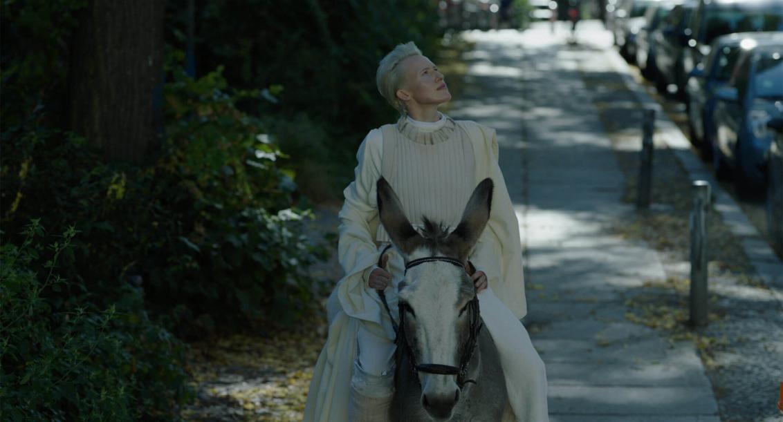 A white-clad, platinum-blonde woman rides a donkey in partial shade on a sidewalk, with trees to her left and parked cars to her right. She puts her head back, her gaze goes up to the right