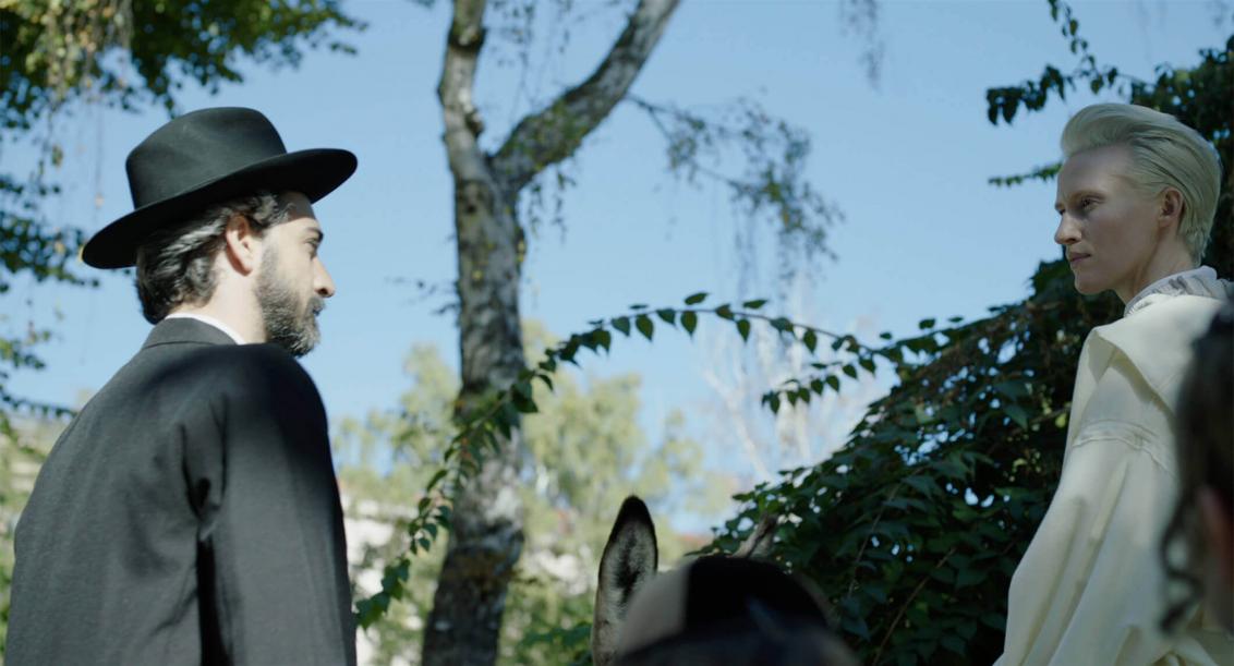 On the right is a short-haired platinum blonde woman riding a donkey, and on the left is a bearded man in a black suit and hat. The two are looking at each other