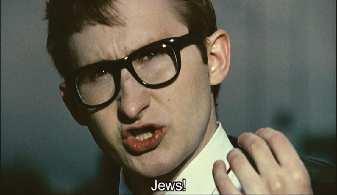 Still from a film showing a close-up of the face of a talking man with short hair and black horn-rimmed glasses, with the subtitle “Jews!”