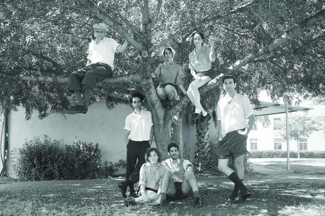 Black and white photograph with young people in summer clothes. Two are sitting in front of a tree in the grass, two are standing under the tree, three are sitting in it. All of them look  into the camera.