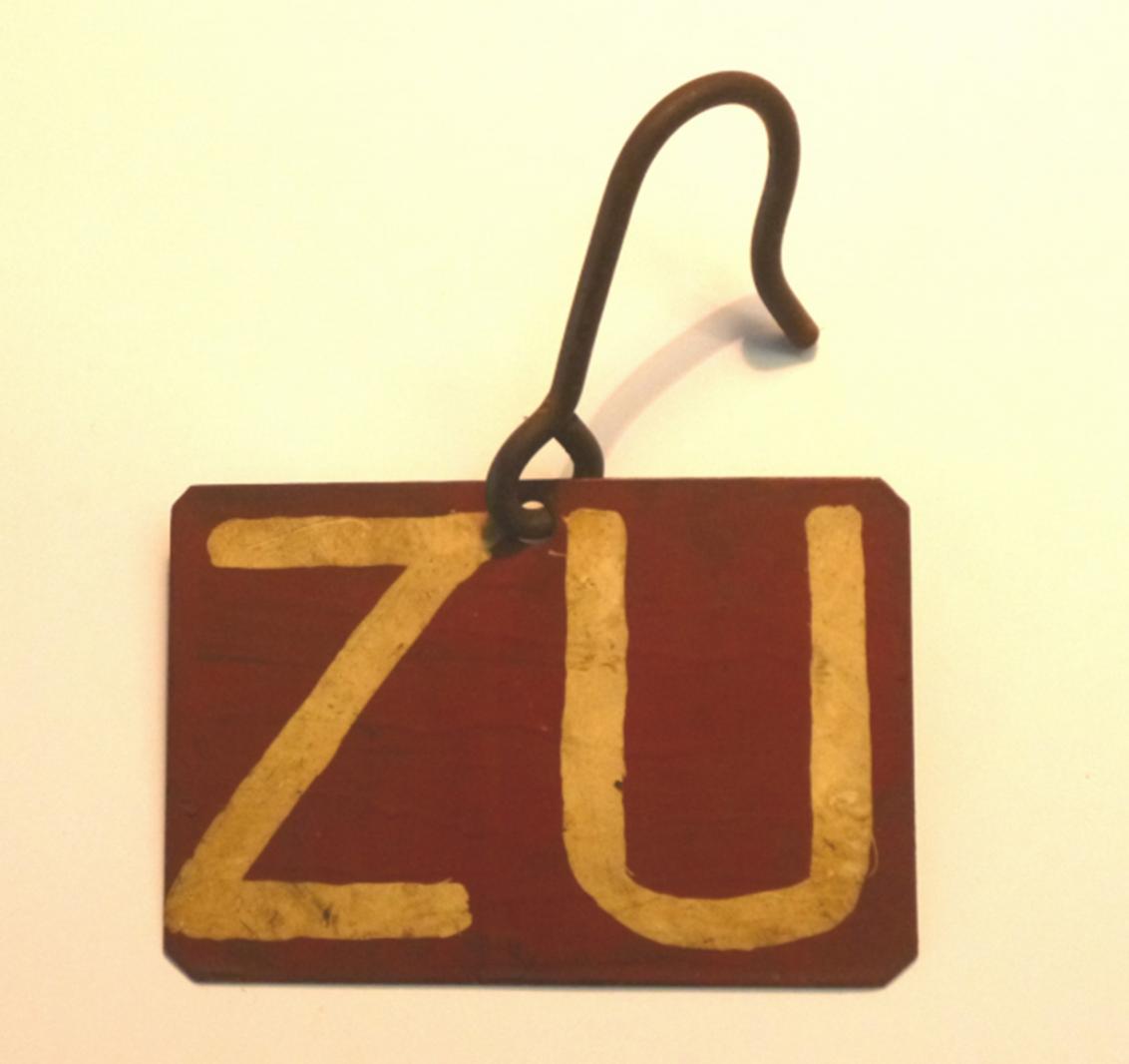 Braunrot in mehreren Schichten lackiertes Metallschild mit der Aufschrift "ZU". Die Ecken des rechteckigen Metallplatte sind abgeschnitten.