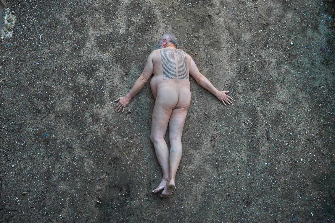 Fotografie eines nackten Mannes, der bäuchlings auf schotteriger Erde liegt, auf seinem Rücken ist zweispaltig eine Passage aus Adornos Minima Moralia tätowiert