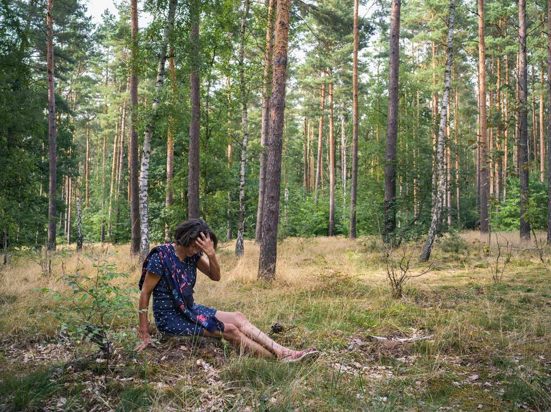 Fotografie einer Frau im blauen Sommerkleid, die im Wald auf dem Boden sitzt und sich über der Stirn in die Haare greift