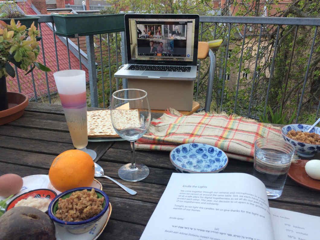 On a wooden table there are various glasses, plates with dishes, bowls and an open book. On a plate lies an orange. Behind the table there is a laptop on a cardboard box, in which a video call can be seen. 