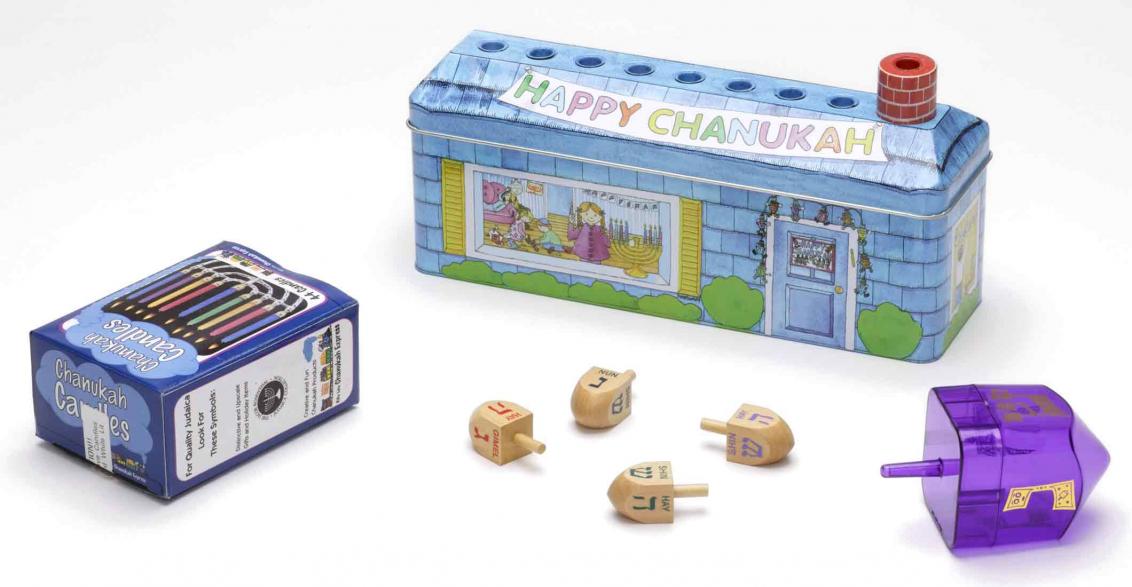 Multi-piece children’s set for Hanukkah consisting of dreidels, candles, and a menorah in the form of a metal box decorated to resemble a house. The roof features eight depressions to hold the candles; the chimney serves as the shamash.