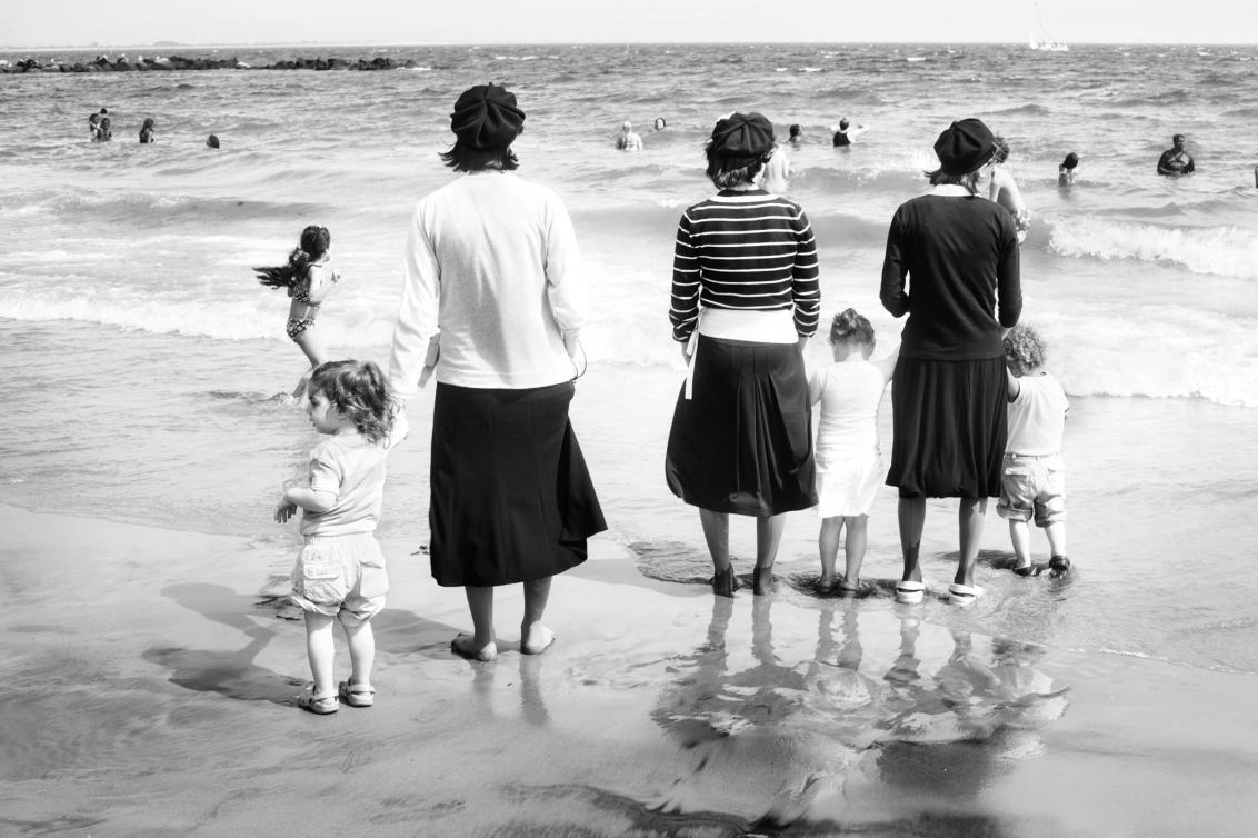 Schwarz-Weiss-Foto von Kleinkindern und orthodoxen Jüdinnen mit Kopfbedeckung und Rock am Strand, von hinten fotografiert