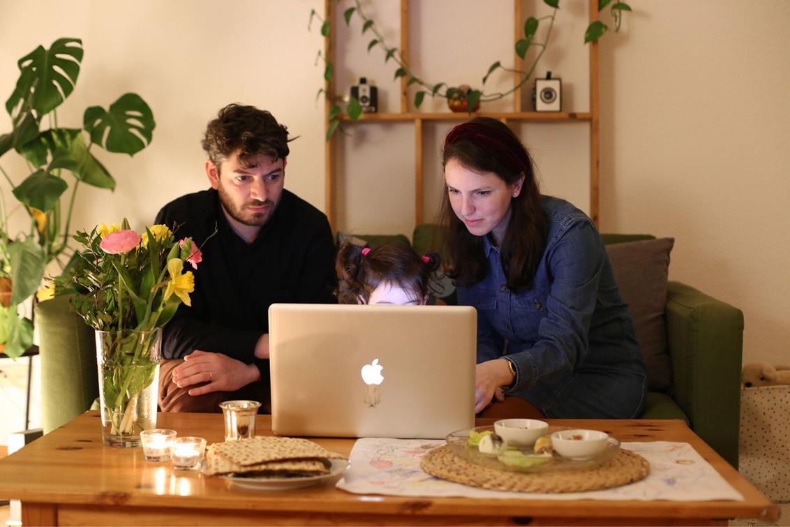 Ein Mann und eine Frau sitzen vor einem Laptop. Über dem Laptop schauen die Haare eines Kindes hervor.