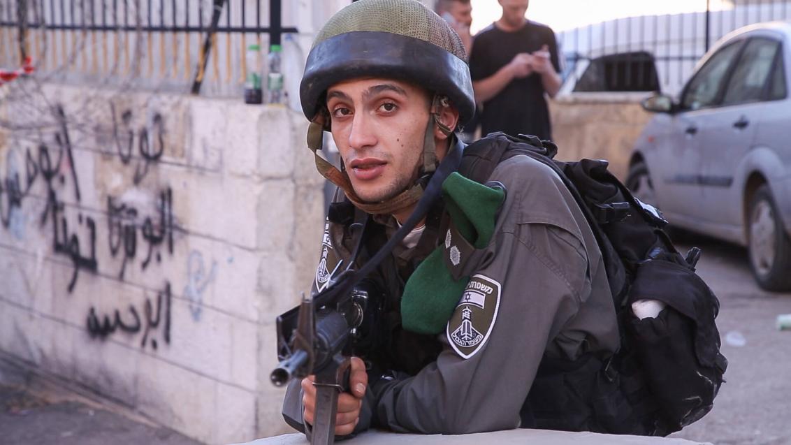 Soldat in Tarnuniform mit Helm und Waffe im Anschlag schaut in die Kamera und zielt Richtung Betrachter*in