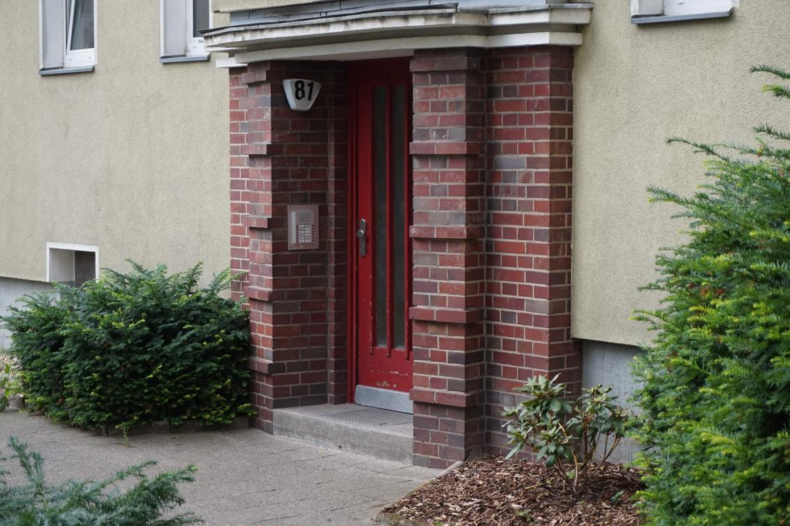 Farbfoto: weinrote Haustür mit Hausnummer „81“, von Backsteinen eingerahmt