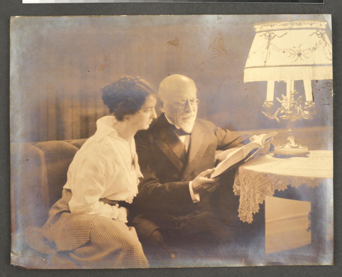 An elderly gentleman with his adult daughter reading under a lampshade.