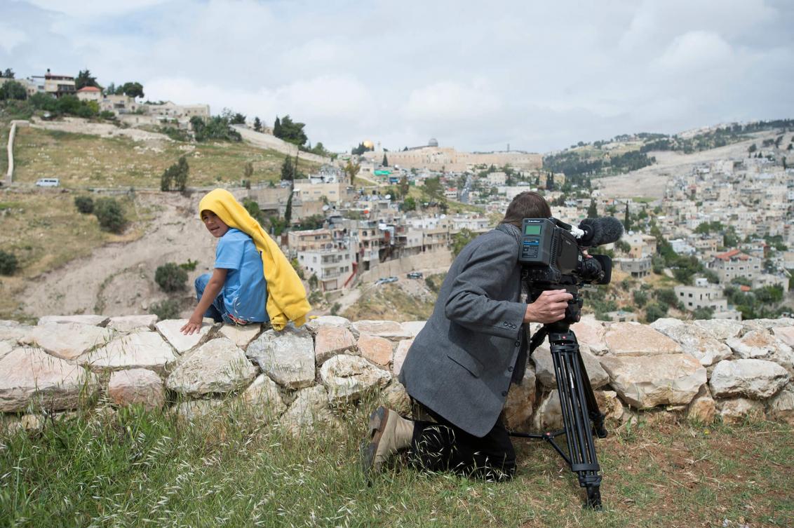 Mann an Filmkamera und Kind, das sich umdreht und in die (Foto-)Kamera schaut, im Bildhintergrund Jerusalem