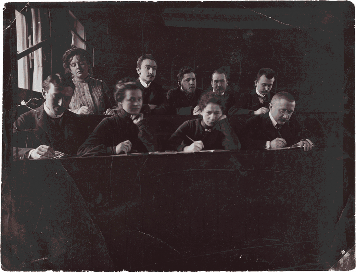 Historische Schwarz-Weiß-Fotografie mehrer schreibender Personen in einem Hörsaal.