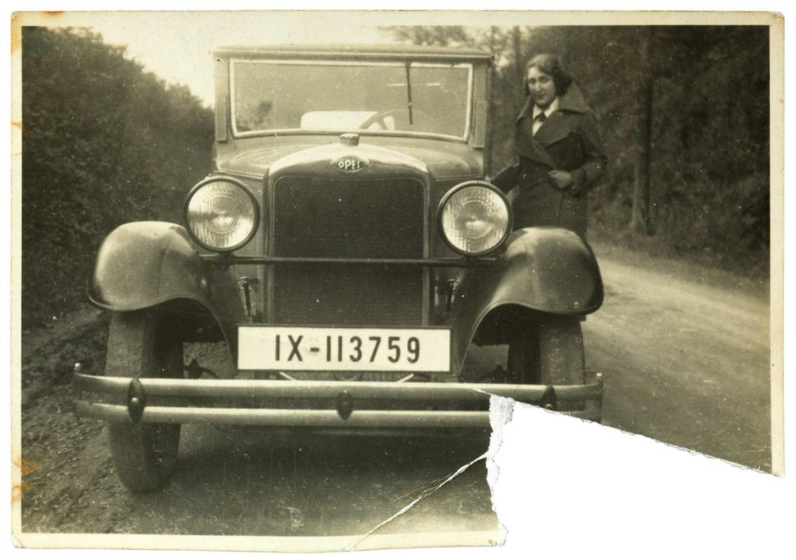 Black and white photo of a young woman in winter clothes next to a car on a country road, the lower right corner of the photograph is torn out.
