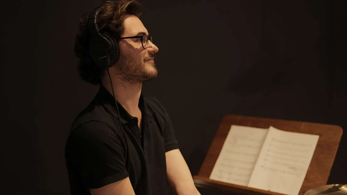 A young man with brunette hair and glasses is wearing headphones, a music stand next to him.