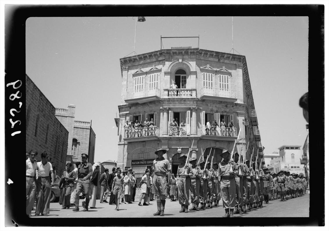 Das Foto in Schwarz-Weiß zeigt eine prunkvolle Fassade mit Balkonen. Darauf stehen Menschen, die auf den Aufmarsch bewaffneter Soldaten in Uniform schauen, der vor dem Gebäude stattfindet.