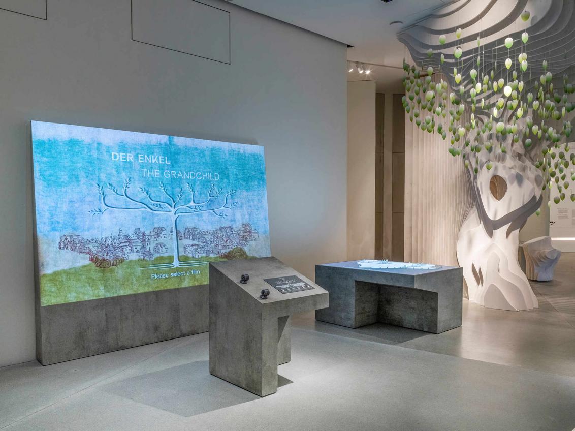 Ausstellungsraum mit Beton-Ausstellungsmöbeln, einer Projektion von einem Baum, um den eine grüne Wiese und Häuser stehen