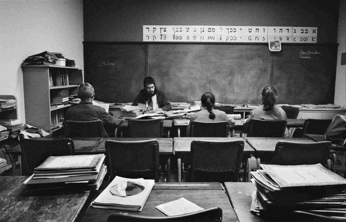 Schwarz-Weiß-Fotographie von einem Klassenraum, Rückenansicht von zwei Schülerinnen und einem Schüler, vor ihnen sitzt ein Lehrer, über der Tafel das hebräische Alphabet