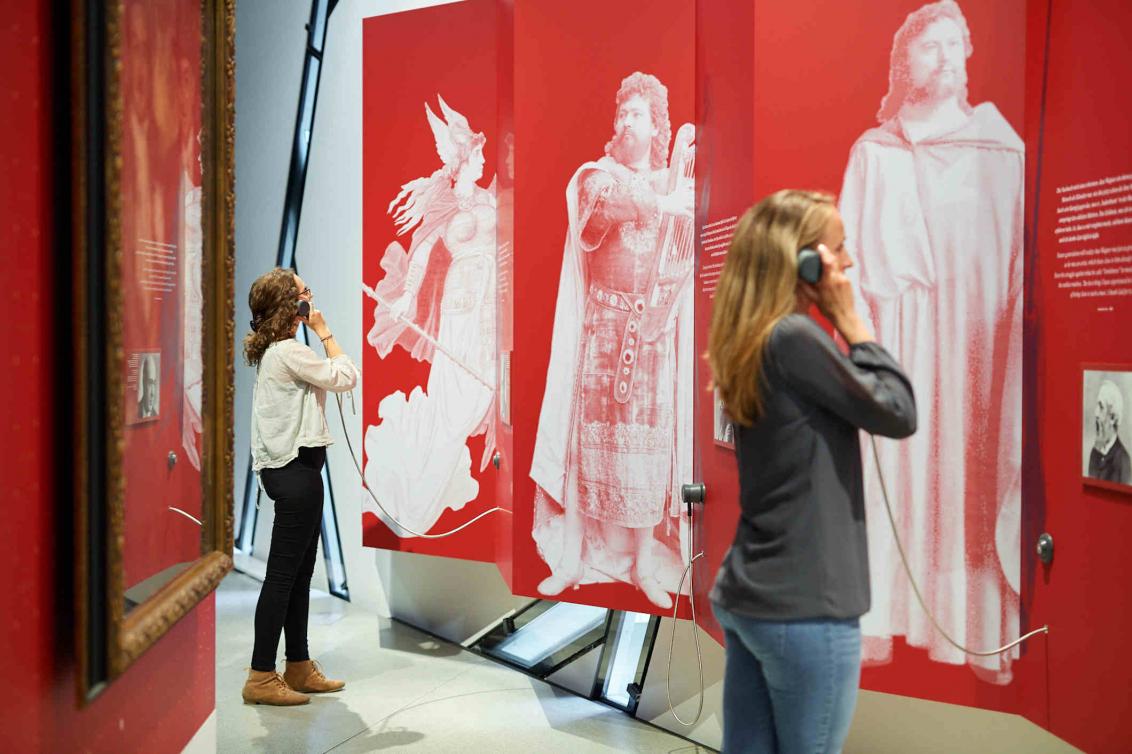 Zwei Besucherinnen stehen mit Hörern in der Hand vor einer roten Wand, auf der weiße Figuren sind
