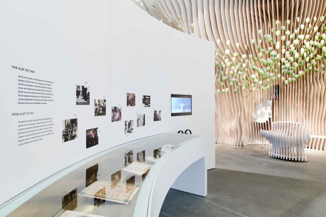 A white curved wall displaying photos and a video, along the length of the wall is a showcase where objects are displayed