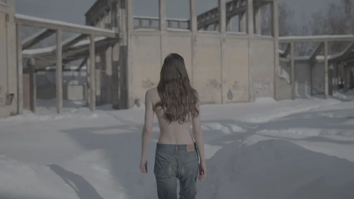 Screenshot of a long-haired person with jeans and bare upper body from behind, walking through snow towards a building.